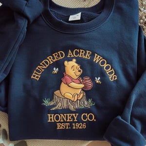 Puede incluir: Sudadera azul marino con dise&ntilde;o bordado de Winnie the Pooh sosteniendo un tarro de miel, sentado en un toc&oacute;n de &aacute;rbol. El texto dice "Hundred Acre Woods Honey Co. Est. 1926". Tambi&eacute;n hay dos abejas bordadas.