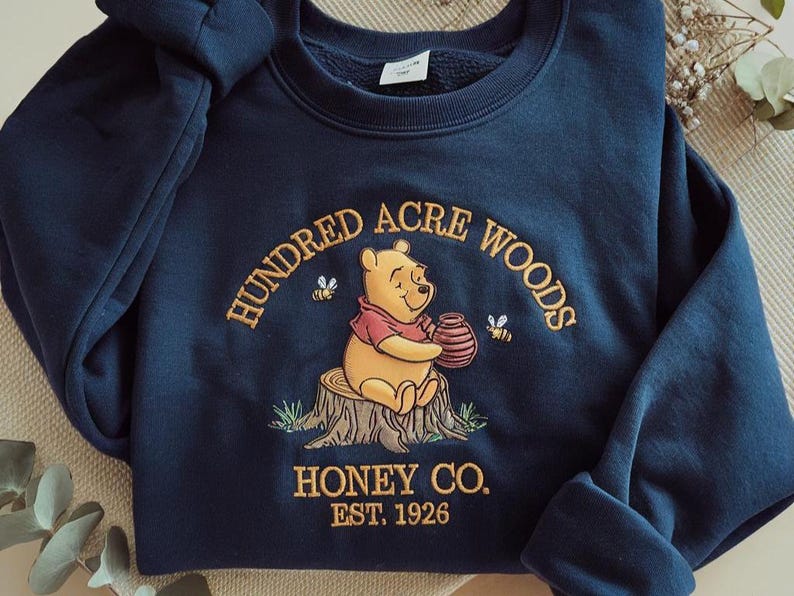 Puede incluir: Sudadera azul marino con dise&ntilde;o bordado de Winnie the Pooh sosteniendo un tarro de miel, sentado en un toc&oacute;n de &aacute;rbol. El texto dice "Hundred Acre Woods Honey Co. Est. 1926". Tambi&eacute;n hay dos abejas bordadas.