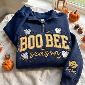 Può includere: Una felpa blu navy con mezza zip caratterizzata da un ricamo giallo e bianco con la scritta BOO BEE season. Il design include illustrazioni di api fantasma, ragnatele e pipistrelli. Il nome Sophia è ricamato sulla manica in corsivo giallo.