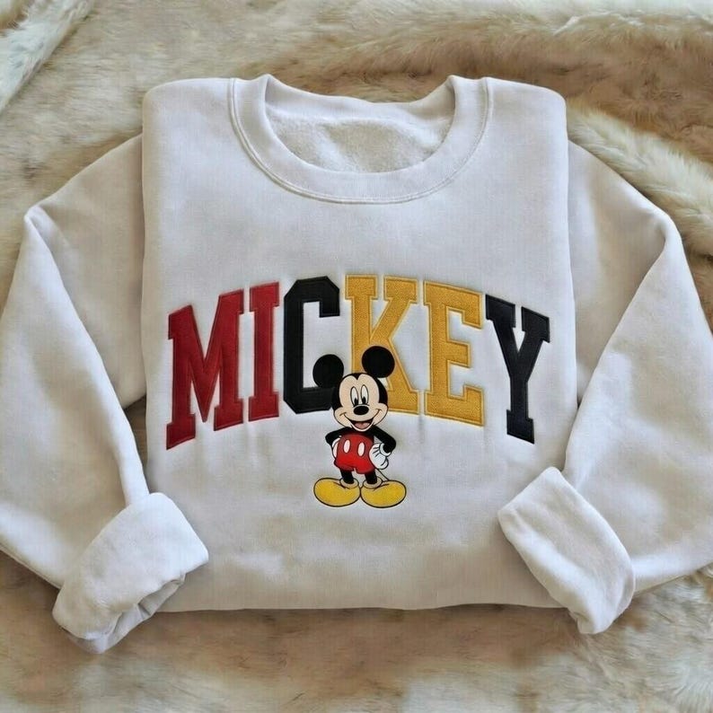 Puede incluir: Sudadera blanca con la palabra "MICKEY" en letras rojas, negras y amarillas. Una imagen de dibujos animados de Mickey Mouse est&aacute; debajo del texto. La sudadera tiene cuello redondo y pu&ntilde;os doblados.