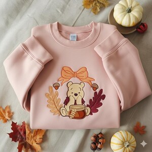 Puede incluir: Sudadera rosa claro con un dise&ntilde;o bordado de un oso de dibujos animados, un lazo, hojas de oto&ntilde;o y bellotas. El oso sostiene un tarro de miel. La sudadera tiene mangas largas y cuello redondo.