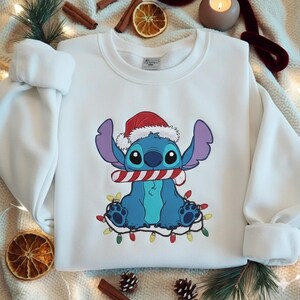May include: White sweatshirt featuring a cartoon character wearing a Santa hat and holding a candy cane. The character is blue with large ears and is surrounded by a string of colorful Christmas lights. The sweatshirt has long sleeves.