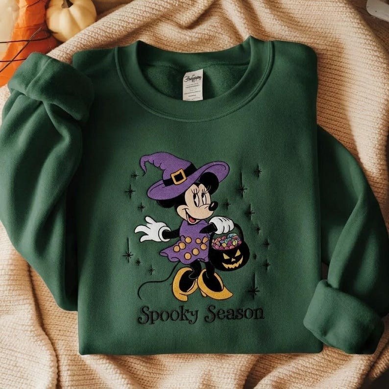 Puede incluir: Una sudadera de manga larga de color verde oscuro con un bordado de un rat&oacute;n de dibujos animados con sombrero y vestido de bruja morados, sosteniendo un cubo de calabaza lleno de dulces. El texto Spooky Season est&aacute; bordado debajo.
