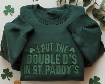 May include: A dark green sweatshirt with the embroidered text "I PUT THE DOUBLE D'S IN ST. PADDY'S". The design includes shamrocks. Gold coins and shamrock cutouts are scattered around the sweatshirt.