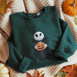 May include: A dark green crewneck sweatshirt featuring an embroidered graphic of a skeletal character with a white skull face and black suit holding an orange jack-o'-lantern pumpkin. The design is centered on the chest of the long-sleeve garment.