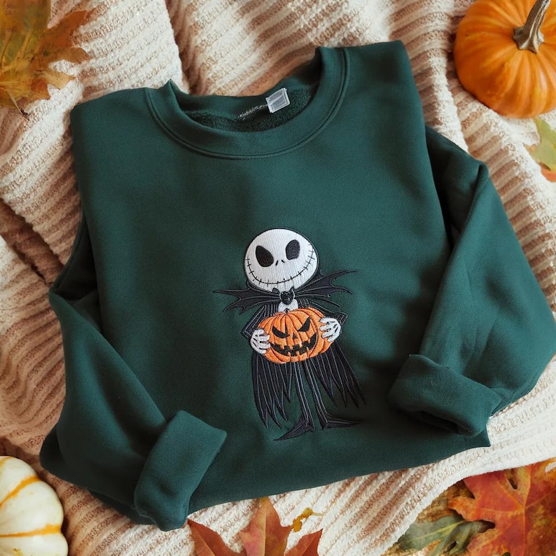 May include: A dark green crewneck sweatshirt featuring an embroidered graphic of a skeletal character with a white skull face and black suit holding an orange jack-o'-lantern pumpkin. The design is centered on the chest of the long-sleeve garment.