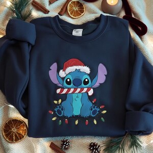 May include: Navy blue sweatshirt featuring a cartoon character wearing a Santa hat and holding a candy cane. The character is surrounded by a string of colorful Christmas lights. The sweatshirt is a cozy, festive garment for the holiday season.