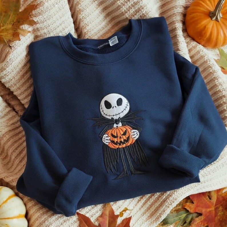 May include: A navy blue crewneck sweatshirt featuring an embroidered graphic of a skeletal character with a white skull face and black suit holding an orange jack-o'-lantern pumpkin. The design is centered on the chest of the long-sleeved garment.