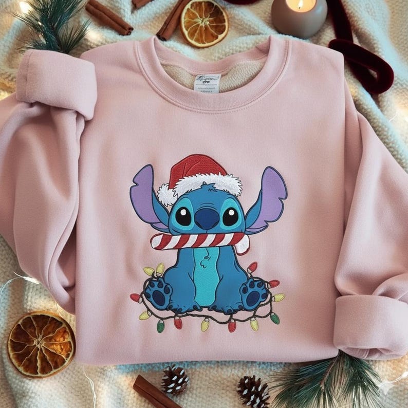 May include: A light pink sweatshirt featuring a cartoon character wearing a Santa hat and holding a candy cane. The character is surrounded by Christmas lights. The sweatshirt has long sleeves with rolled cuffs.