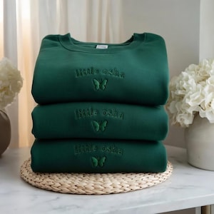 May include: A stack of three dark forest green crewneck sweatshirts. Each sweatshirt features the embroidered text "little osha" in a matching dark green thread, positioned above a small, simple embroidered butterfly icon centered on the chest.