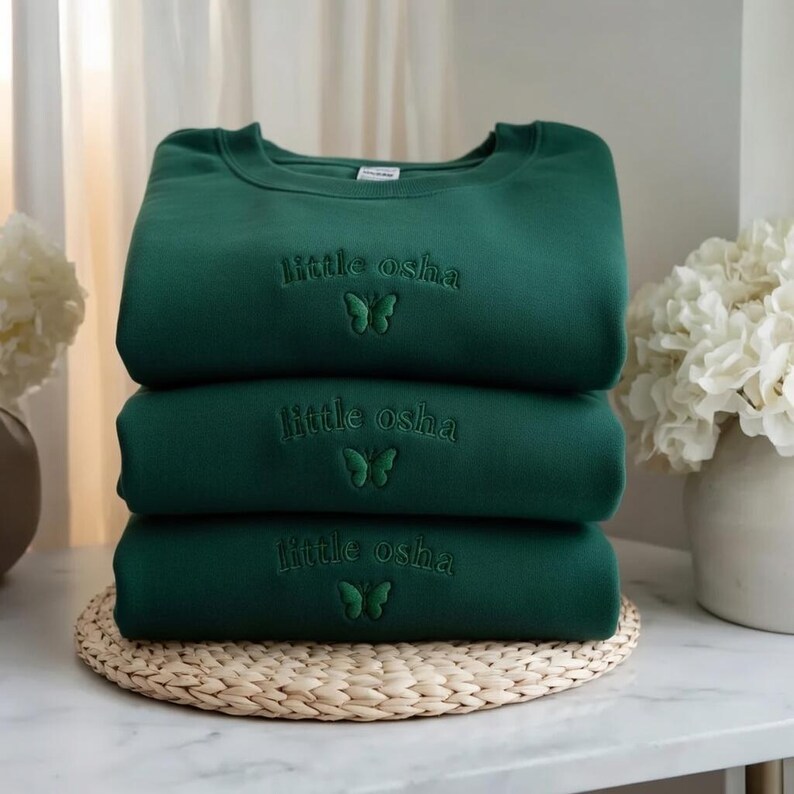 May include: A stack of three dark forest green crewneck sweatshirts. Each sweatshirt features the embroidered text "little osha" in a matching dark green thread, positioned above a small, simple embroidered butterfly icon centered on the chest.