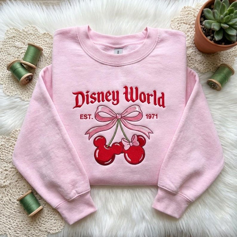 May include: A light pink sweatshirt with the words "Disney World" embroidered in red script. Below the text is a pink bow and two red cherry-shaped Mickey Mouse heads. The text "EST. 1971" is also embroidered on the sweatshirt.