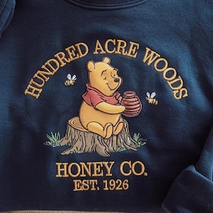Puede incluir: Sudadera azul marino con un bordado de Winnie the Pooh sosteniendo un tarro de miel. El texto "Hundred Acre Woods" y "Honey Co. Est. 1926" est&aacute;n bordados en dorado. La imagen presenta dos abejas.