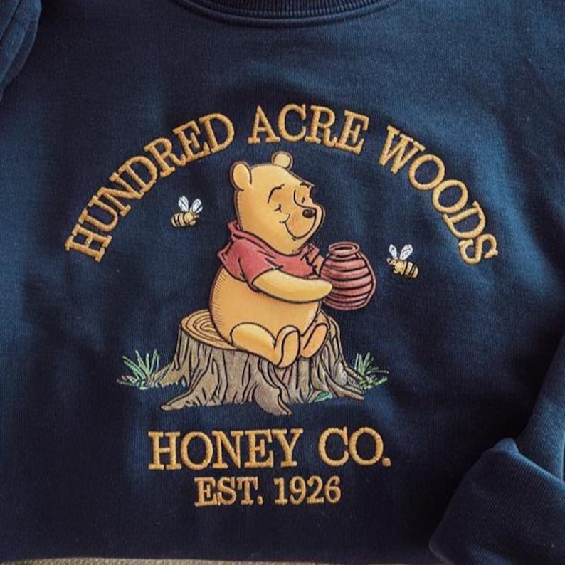 Puede incluir: Sudadera azul marino con un bordado de Winnie the Pooh sosteniendo un tarro de miel. El texto "Hundred Acre Woods" y "Honey Co. Est. 1926" est&aacute;n bordados en dorado. La imagen presenta dos abejas.