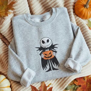 May include: A light gray crewneck sweatshirt featuring an embroidered graphic of a skeletal character with a round white head and black facial features, wearing a black coat and holding a bright orange carved pumpkin with a jagged mouth.