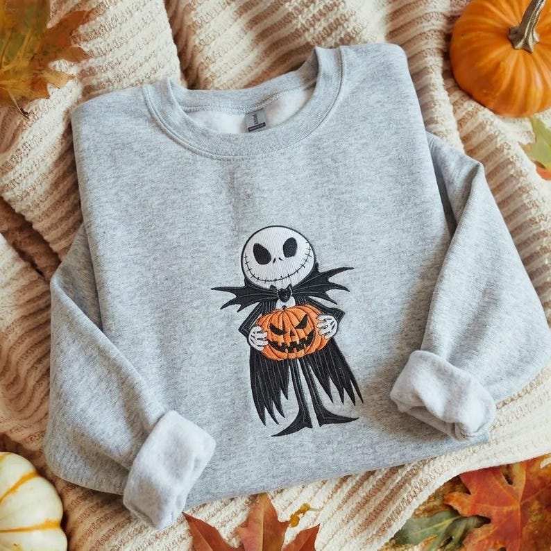 May include: A light gray crewneck sweatshirt featuring an embroidered graphic of a skeletal character with a round white head and black facial features, wearing a black coat and holding a bright orange carved pumpkin with a jagged mouth.