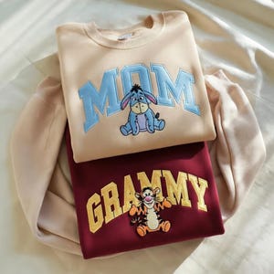 May include: Two sweatshirts, one tan and one burgundy, are stacked. The tan sweatshirt has the word "MOM" in blue block letters with a cartoon donkey. The burgundy sweatshirt has the word "GRAMMY" in gold block letters with a cartoon tiger.