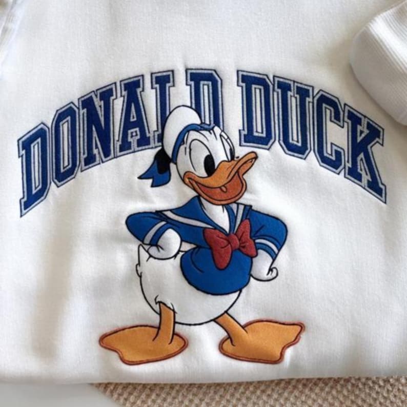 May include: White sweatshirt featuring a cartoon image of Donald Duck in a sailor outfit. The text "DONALD DUCK" is arched above the character in blue block letters. The design includes a red bow tie and a blue sailor suit.