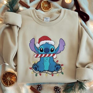 May include: Beige sweatshirt featuring a blue cartoon character wearing a Santa hat and holding a candy cane. The character is surrounded by a string of colorful Christmas lights. The sweatshirt has long sleeves and a crew neck.