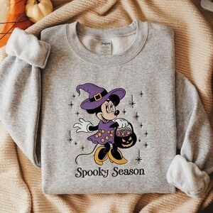 Puede incluir: Una sudadera gris jaspeado de cuello redondo con el gr&aacute;fico de un rat&oacute;n de dibujos animados con sombrero y vestido de bruja morado, sosteniendo un cubo de calabaza lleno de dulces. El texto Spooky Season est&aacute; impreso en negro debajo.