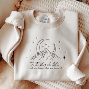 Pode incluir: Uma camisola de gola redonda cor de creme com bordado preto de uma lua crescente, estrelas e picos de montanhas. Abaixo do design, o texto diz: To the stars who listen and the dreams that are answered. A peça tem mangas compridas.