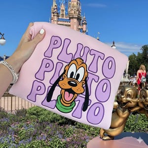 Puede incluir: Una sudadera rosa claro con Pluto de Disney. La palabra "Pluto" está impresa en letras grandes de color lavanda. La cara de Pluto está en el centro, con la lengua fuera. El fondo incluye un castillo y una estatua.