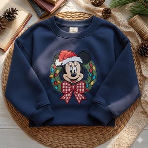 May include: Navy blue sweatshirt featuring a Minnie Mouse design. The design includes Minnie wearing a Santa hat, surrounded by a Christmas wreath with colorful lights and a red and white checkered bow. The sweatshirt is a crew neck style.