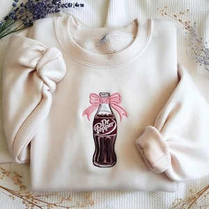 Op de afbeelding: Crèmekleurige sweatshirt met een ronde hals en lange mouwen. De voorkant heeft een geborduurde Dr. Pepper-fles met een roze strik. Het Dr. Pepper-logo is zichtbaar. De sweatshirt is gemaakt van een zacht materiaal.
