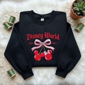 May include: Black crewneck sweatshirt with red embroidered "Disney World" text and a Mickey and Minnie Mouse cherry design. The sweatshirt also features the text "EST. 1971".