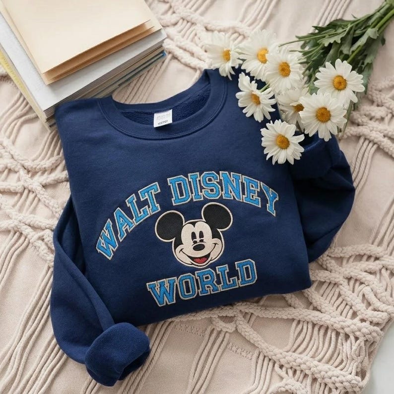 Puede incluir: Una sudadera azul marino de cuello redondo con el texto WALT DISNEY WORLD en fuente azul claro con borde blanco. Centrado entre las palabras hay un gr&aacute;fico de la cara de Mickey Mouse. La sudadera tiene mangas largas y cuello acanalado.