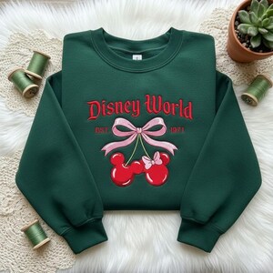 May include: A dark green sweatshirt with the words "Disney World" embroidered in red script. Below the text is a pink bow and two red Mickey Mouse-shaped cherries. The year "1971" is embroidered below the bow.