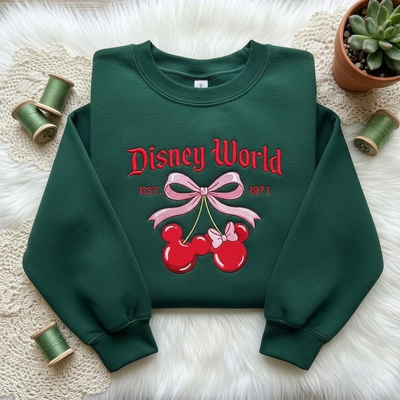 May include: A dark green sweatshirt with the words "Disney World" embroidered in red script. Below the text is a pink bow and two red Mickey Mouse-shaped cherries. The year "1971" is embroidered below the bow.
