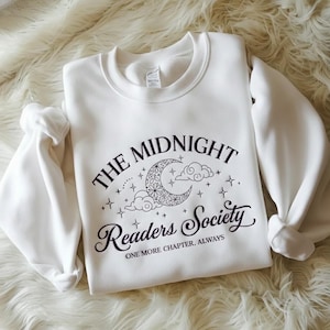Op de afbeelding: Witte sweatshirt met de tekst "THE MIDNIGHT Readers Society" in zwart schrift. Het ontwerp bevat een maan, wolken en sterren. De zin "ONE MORE CHAPTER, ALWAYS" staat eronder gedrukt. De sweatshirt heeft lange mouwen.