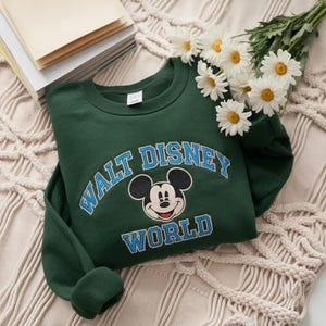 Puede incluir: Una sudadera de cuello redondo de color verde oscuro con un gr&aacute;fico de Mickey Mouse centrado entre las palabras WALT DISNEY y WORLD. El texto est&aacute; impreso en una fuente azul claro con un borde blanco fino, y la sudadera tiene mangas largas.