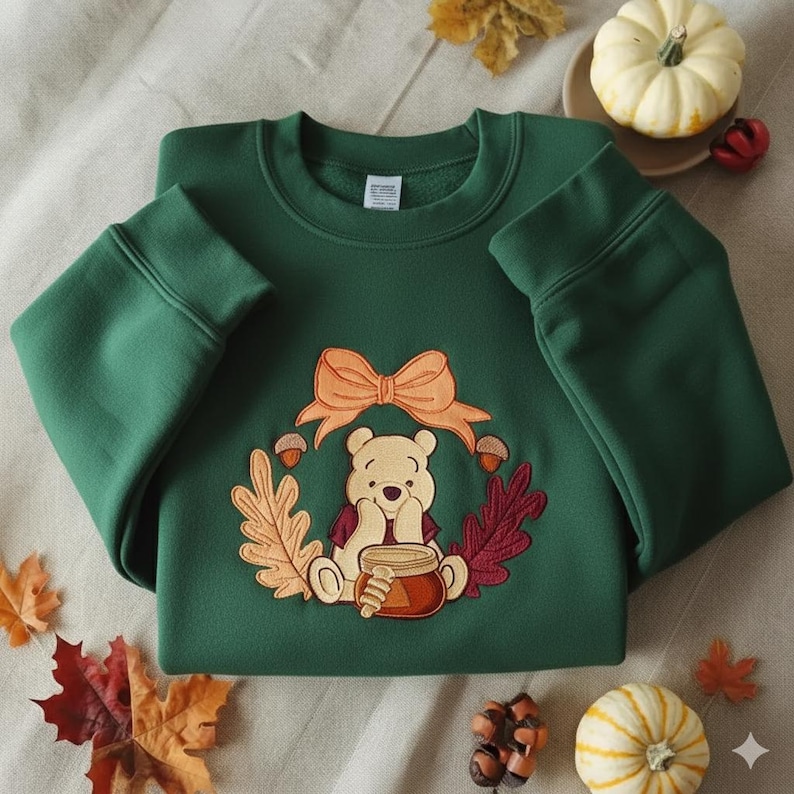 Puede incluir: Sudadera verde oscuro con un dise&ntilde;o bordado de un oso sosteniendo un tarro de miel, rodeado por una corona de hojas de oto&ntilde;o, bellotas y un lazo. La sudadera est&aacute; doblada, con las mangas visibles.