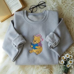 May include: A light gray sweatshirt featuring a cartoon illustration of Winnie the Pooh reading a book. Pooh is wearing a pink nightcap and purple shirt. The sweatshirt has a crew neck and long sleeves with rolled cuffs. A book and glasses are in the background.
