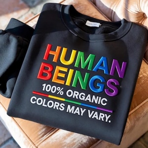 May include: Black sweatshirt with the embroidered text "HUMAN BEINGS 100% ORGANIC COLORS MAY VARY" in rainbow colors. The sweatshirt is folded on a light brown surface.