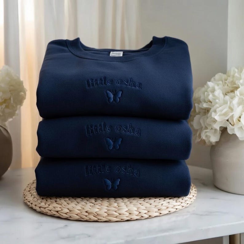 May include: A stack of three navy blue crewneck sweatshirts, each featuring the embroidered text "little osha" in a matching dark blue thread, positioned above a small, detailed butterfly icon centered on the chest area of the garment.