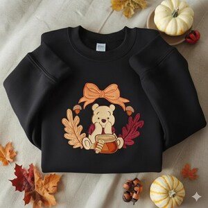 Puede incluir: Sudadera negra con un dise&ntilde;o bordado de Winnie the Pooh. El dise&ntilde;o incluye un oso, un tarro de miel, un lazo, bellotas y hojas de oto&ntilde;o en tonos naranja, rojo y marr&oacute;n. Decoraciones oto&ntilde;ales.