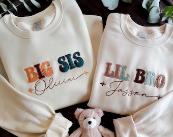 May include: Two cream-colored sweatshirts with embroidered text. One says "BIG SIS" and the other says "LIL BRO". Both have names embroidered below the text. A small teddy bear sits in front of the sweatshirts.