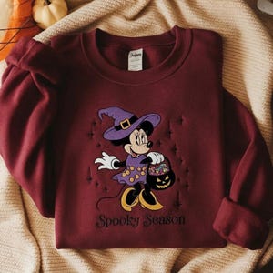 Puede incluir: Una sudadera de cuello redondo color granate con un bordado de un rat&oacute;n de dibujos animados con sombrero y vestido de bruja morado, sosteniendo un cubo de dulces de calabaza. El texto "Spooky Season" est&aacute; bordado en negro debajo.