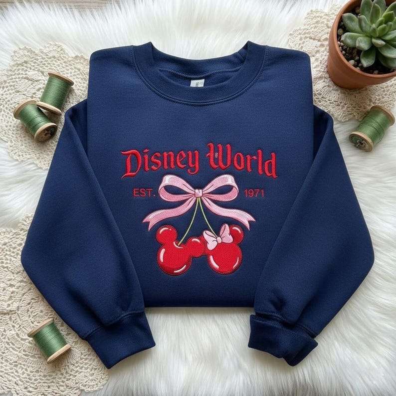 May include: Navy blue sweatshirt with red embroidered "Disney World" text, a pink bow, and cherry-shaped Mickey Mouse heads. The text "EST. 1971" is also embroidered. The sweatshirt is a casual style.