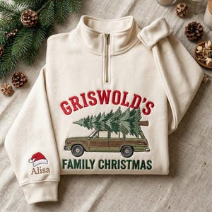 Puede incluir: Una sudadera color crema con cremallera corta y el texto bordado Griswold's Family Christmas sobre un coche familiar verde que transporta un pino. La manga incluye un gorro de Papá Noel bordado y el nombre Alisa.