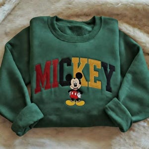Puede incluir: Una sudadera verde con la palabra "MICKEY" bordada en rojo, negro y amarillo, y una imagen bordada de Mickey Mouse. La sudadera tiene cuello redondo y mangas largas.