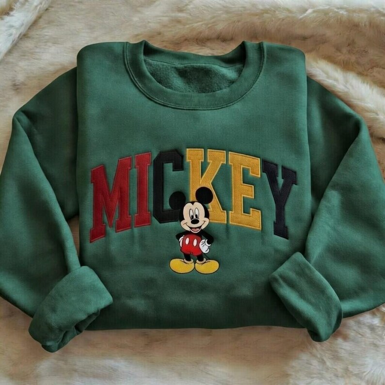 Puede incluir: Una sudadera verde con la palabra "MICKEY" bordada en rojo, negro y amarillo, y una imagen bordada de Mickey Mouse. La sudadera tiene cuello redondo y mangas largas.