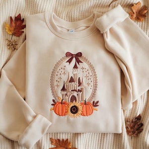 May include: A cream-coloured crewneck sweatshirt featuring an embroidered design of a castle inside an oval frame topped with a bow. Below the castle are two orange pumpkins and a central sunflower, all rendered in warm autumn-toned embroidery thread.