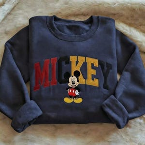 Puede incluir: Sudadera azul marino con la palabra "MICKEY" en letras de bloque rojas, negras y amarillas. Un dibujo animado de Mickey Mouse est&aacute; bordado debajo del texto. La sudadera tiene mangas largas y cuello redondo.
