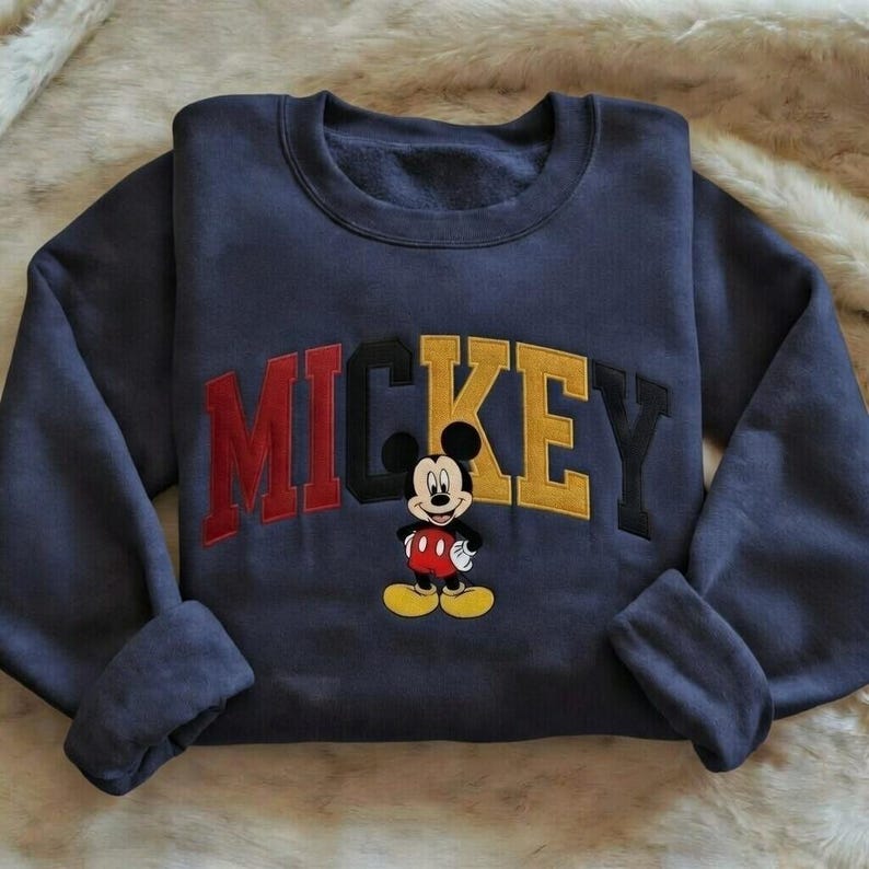 Puede incluir: Sudadera azul marino con la palabra "MICKEY" en letras de bloque rojas, negras y amarillas. Un dibujo animado de Mickey Mouse est&aacute; bordado debajo del texto. La sudadera tiene mangas largas y cuello redondo.