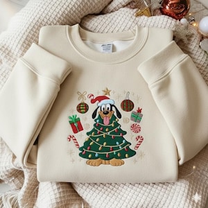 Puede incluir: Sudadera de color crema con un diseño navideño bordado. El diseño presenta un perro de dibujos animados con un gorro de Papá Noel y un árbol de Navidad. El árbol está decorado con luces, adornos y regalos. Bastones de caramelo y estrellas completan el look festivo.