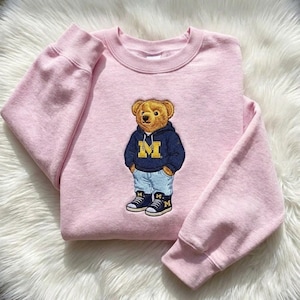 May include: A light pink sweatshirt featuring an embroidered bear wearing a navy blue hoodie with a gold "M" and blue jeans. The bear is also wearing black sneakers with the letter "M" on them.
